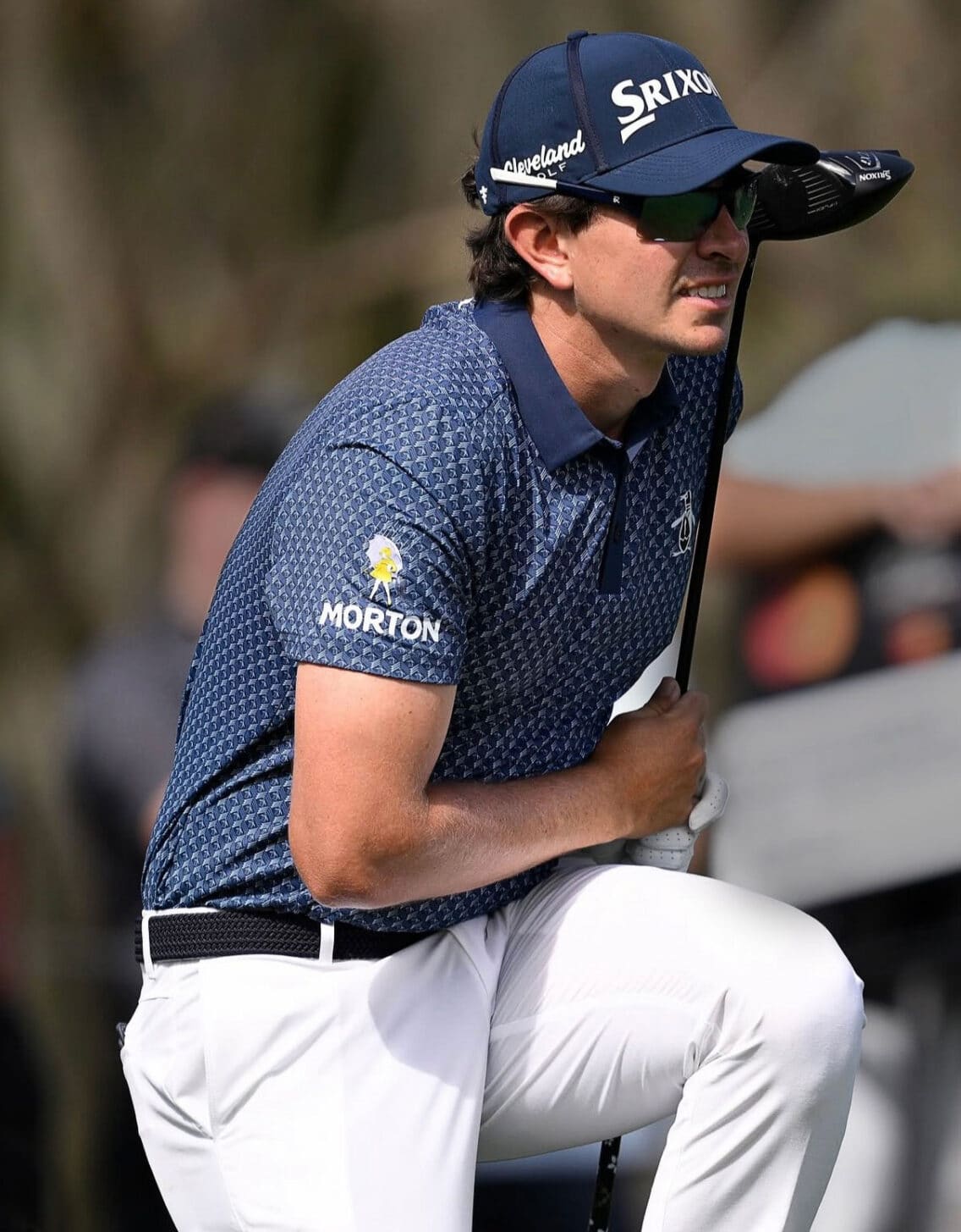 Professional golfer preparing to take a shot, wearing a blue patterned polo shirt and white pants, holding a golf club, with a focused expression. Brand logos visible on the shirt and cap.